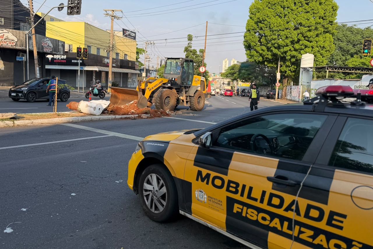 Máquina retroescavadeira corta o canteiro no cruzamento da Rua 240 com a Avenida T-2, no Setor Coimbra, em Goiânia, ao lado de viatura da fiscalização de mobilidade