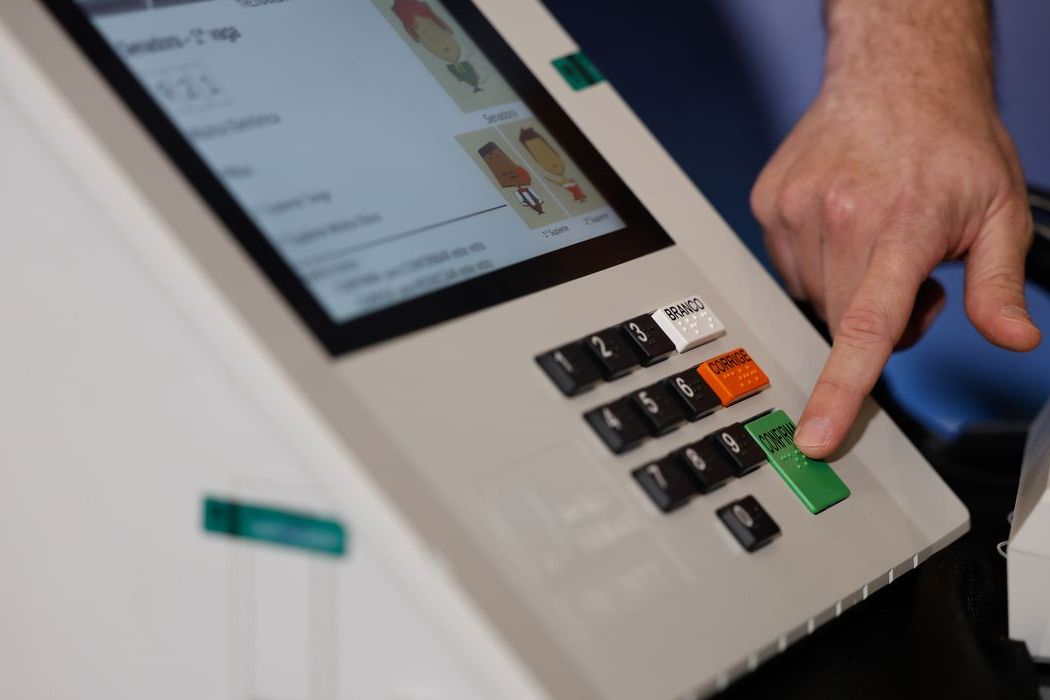 Dedo de um eleitor pressiona a tecla verde Confirma de uma urna eletrônica, com a tela exibindo o painel de votação