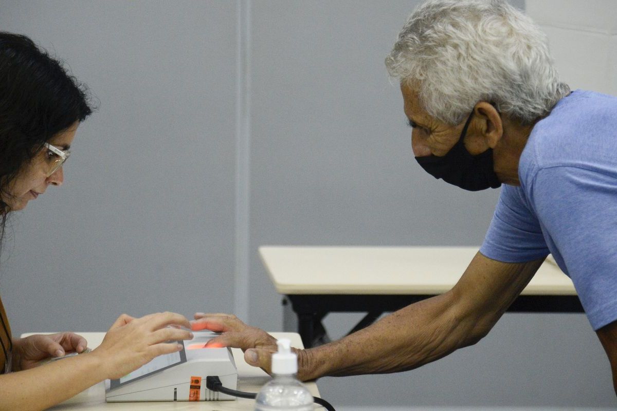 Eleitor idoso apoia a mão no leitor biométrico diante da mesária, em seção eleitoral
