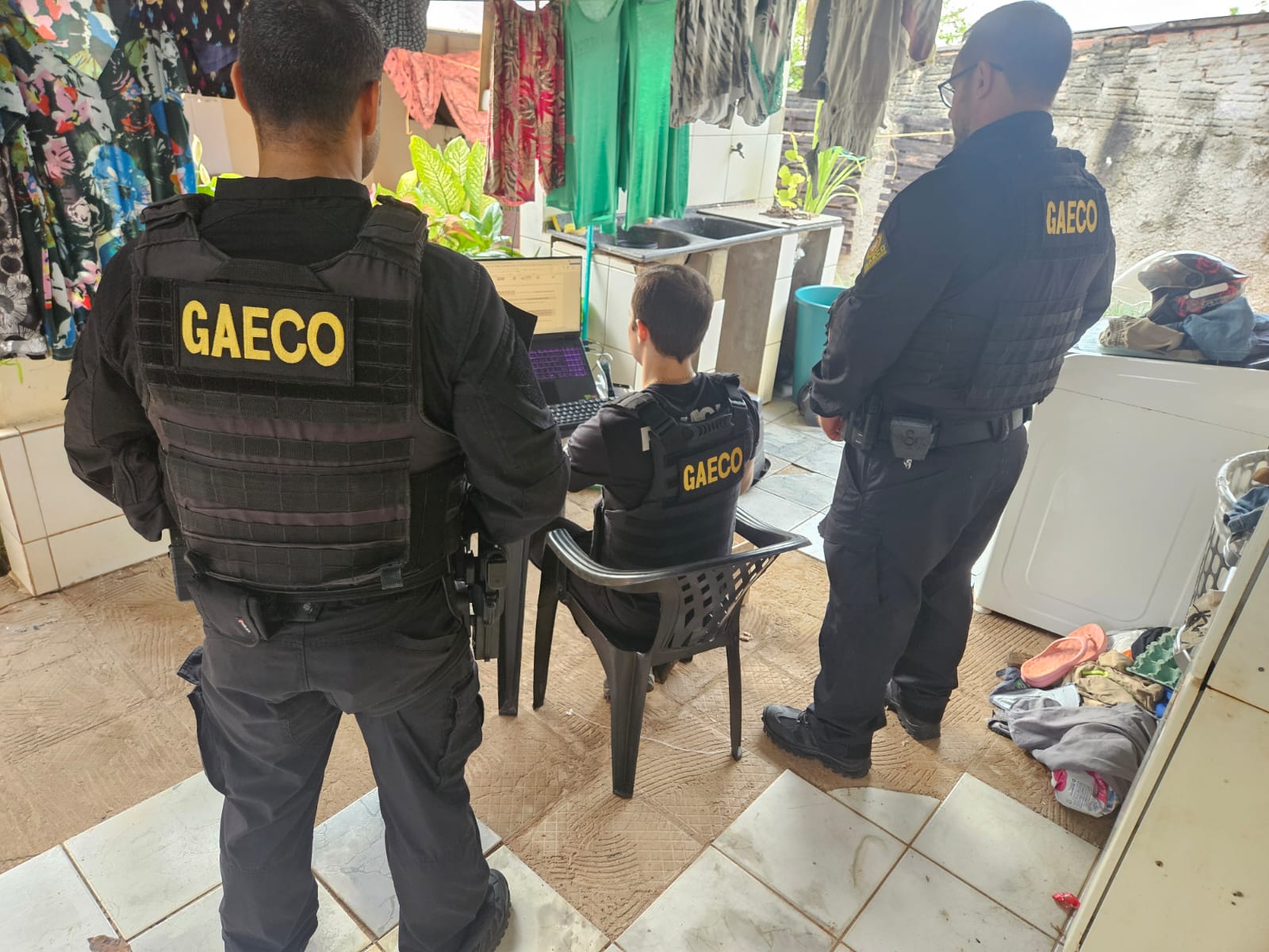 Três agentes com coletes do Gaeco cumprem mandado de busca em residência, um deles diante de um notebook