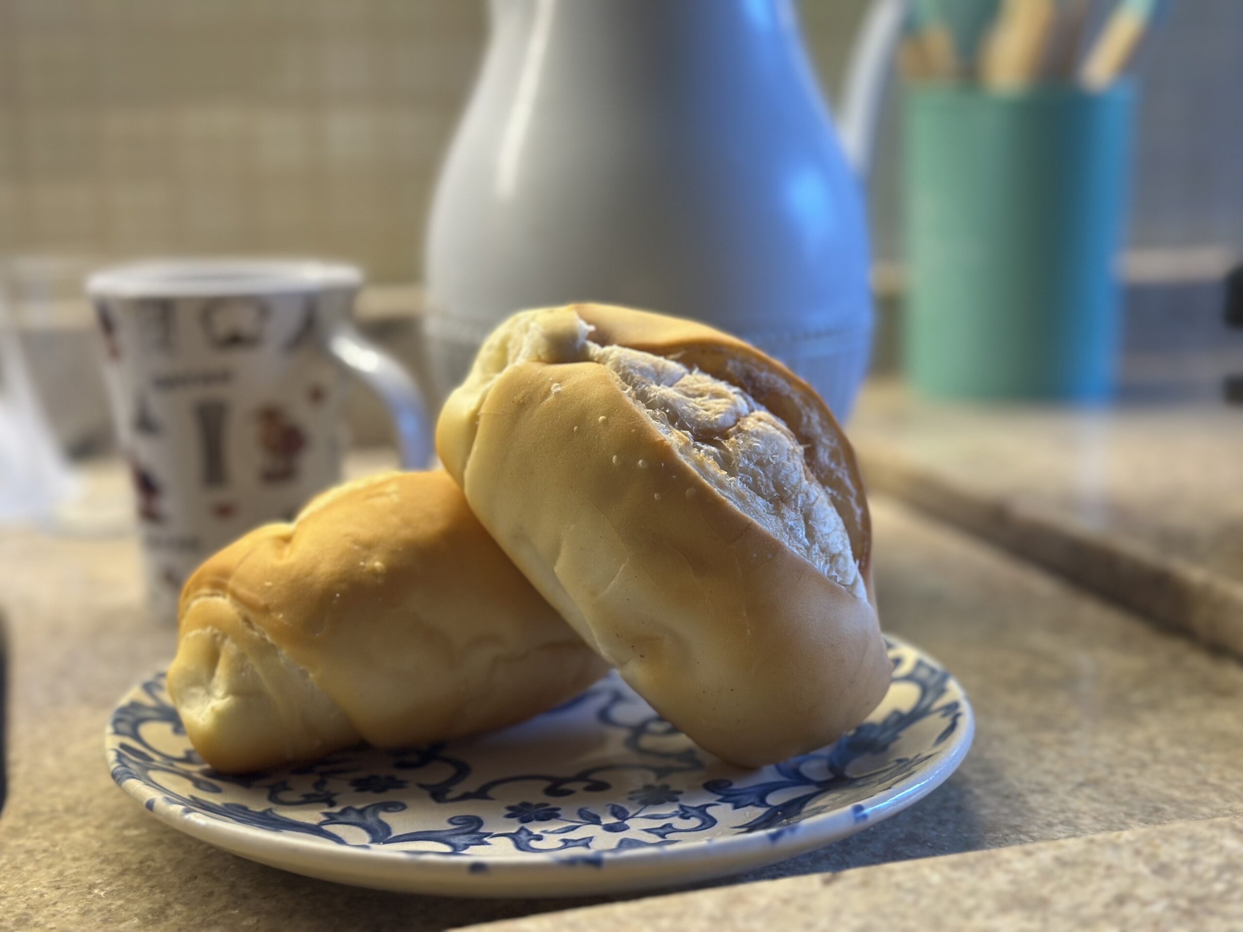 Dois pães franceses em um prato, com xícara e garrafa térmica ao fundo