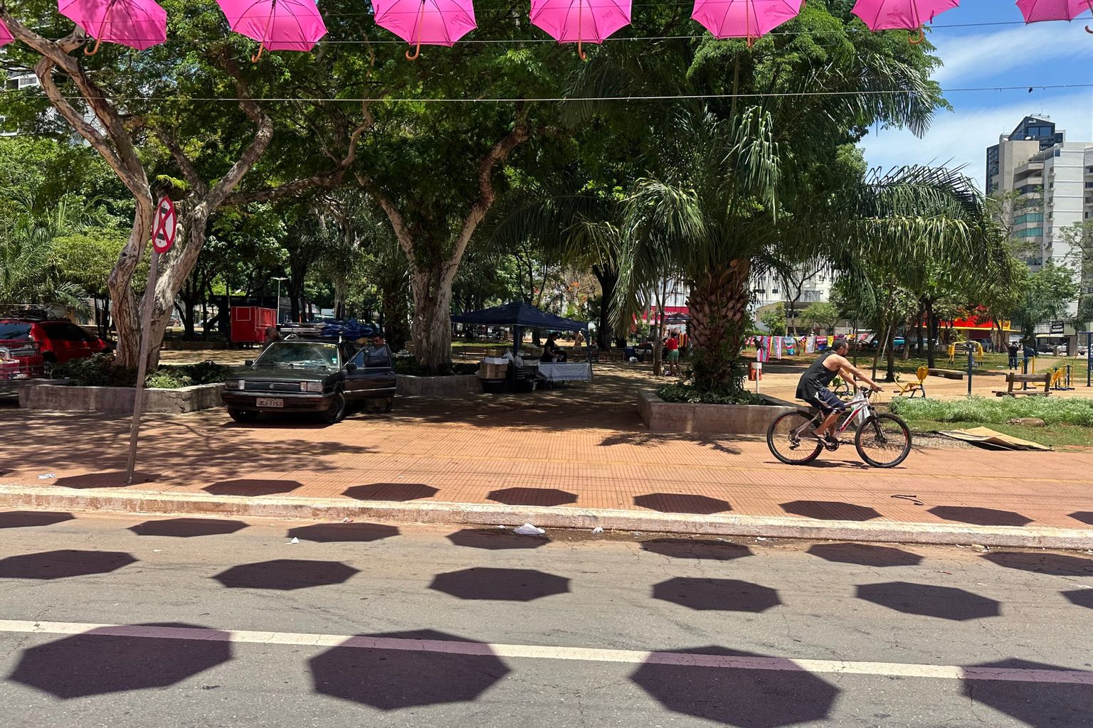 Pra&ccedil;a Tamandar&eacute; arborizada, com guarda-chuvas cor-de-rosa suspensos sobre a via e ambulantes montados ao fundo