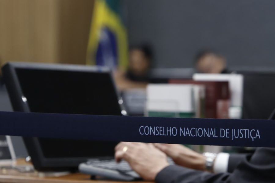 Placa do Conselho Nacional de Justiça sobre a mesa, com computador e a bandeira do Brasil ao fundo