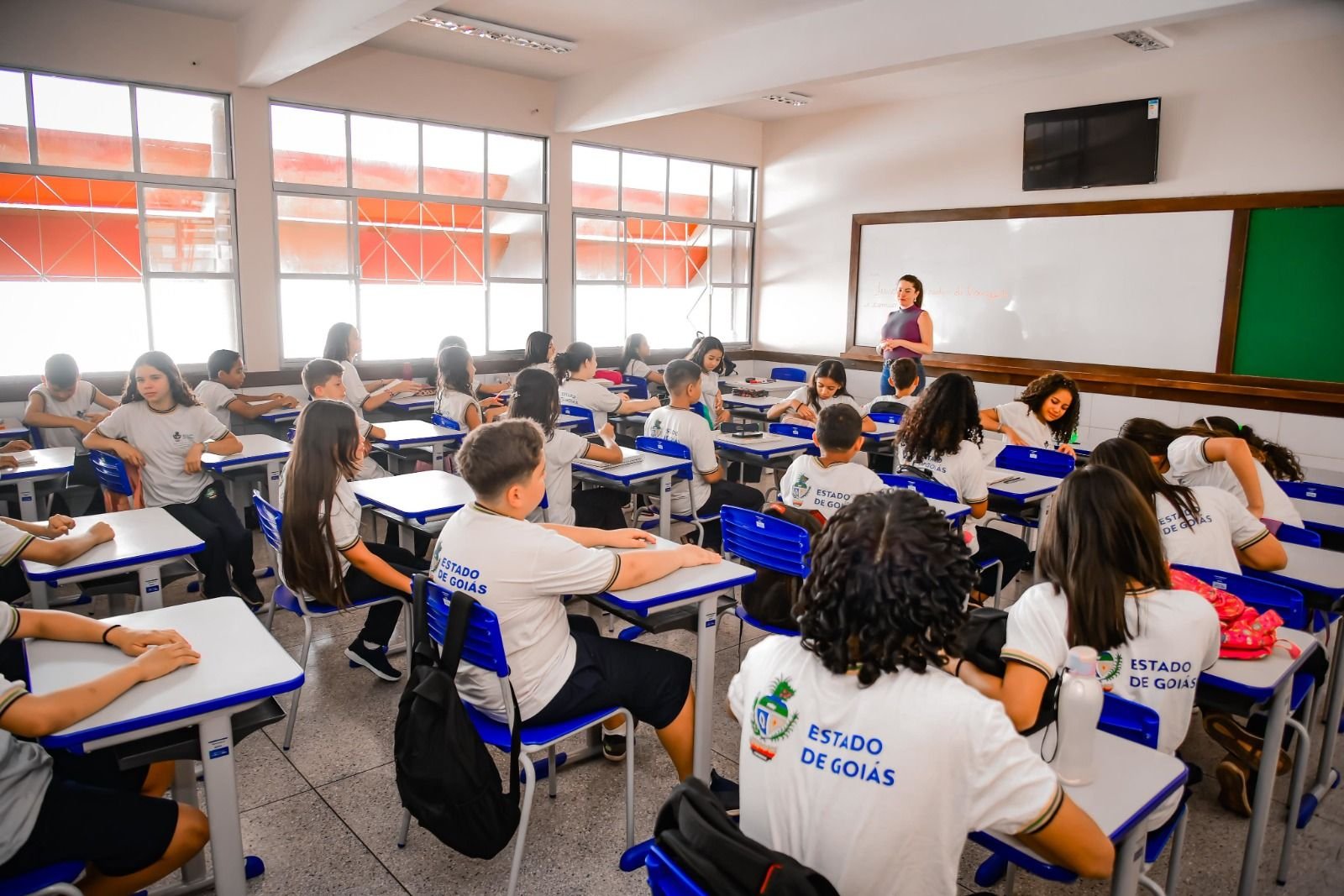 Alunos uniformizados sentados em sala de aula da rede estadual de ensino de Goiás, com a professora ao fundo