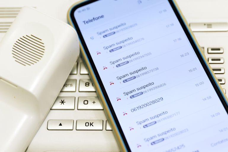 Tela de telefone celular com lista de ligações marcadas como spam suspeito, ao lado de aparelho fixo