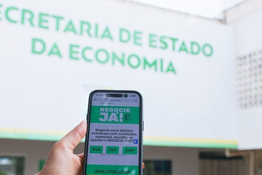 Mão segura celular com a página do Negocie Já II diante da fachada da Secretaria de Estado da Economia