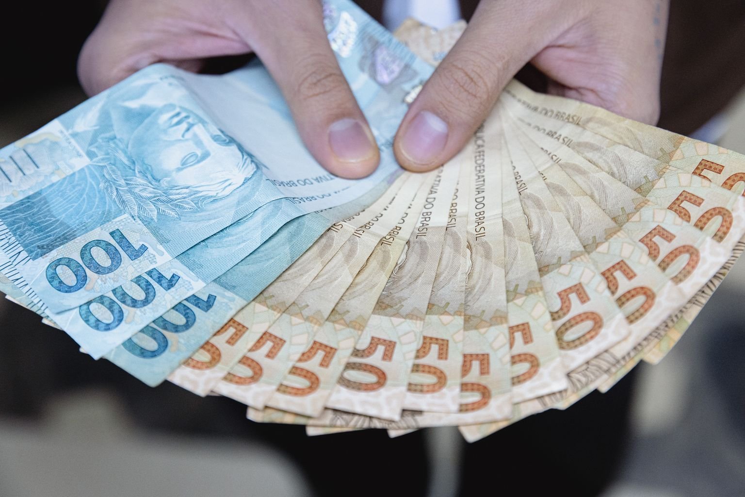 Mãos seguram leque de notas de real de R$ 100 e R$ 50, ilustrando os prêmios da Nota Fiscal Goiana