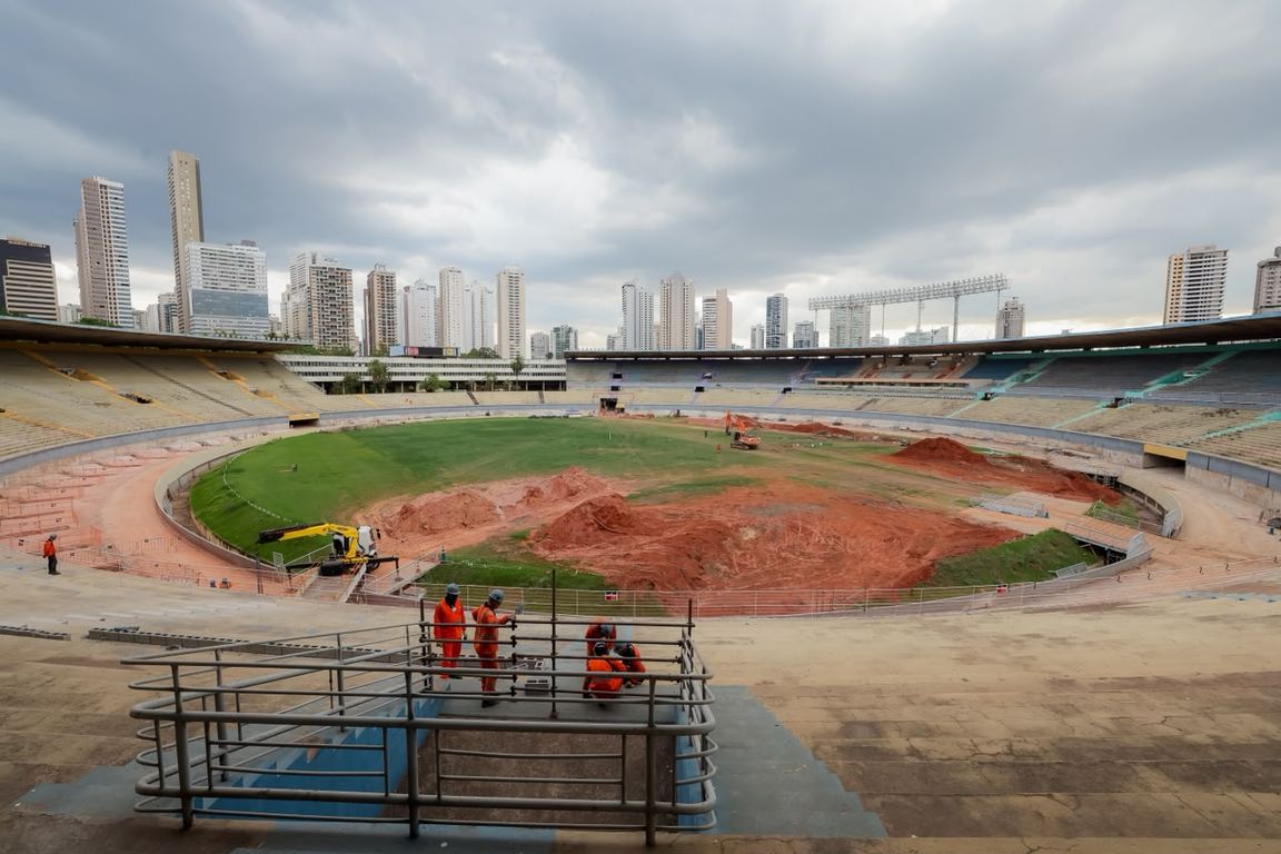 Operários observam a obra no gramado do Estádio Serra Dourada, com prédios de Goiânia ao fundo