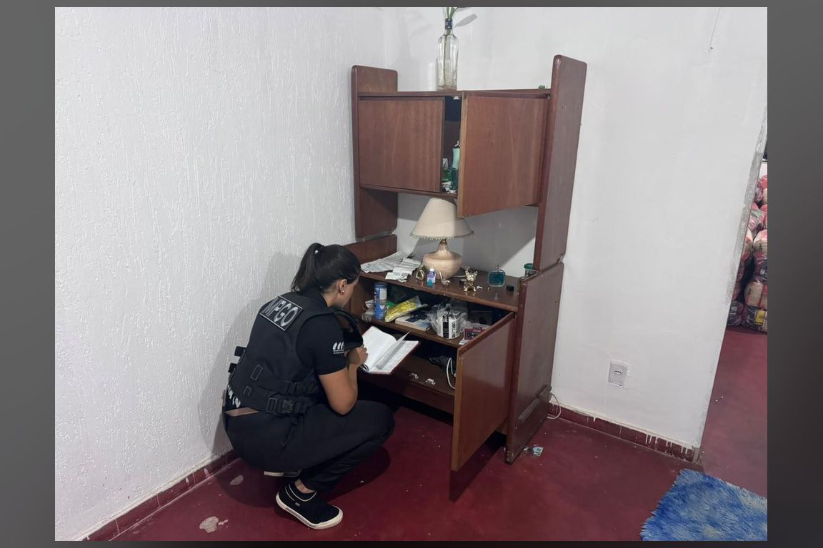 Servidora do Ministério Público de Goiás agachada, examinando documentos em um móvel de madeira durante cumprimento de mandado de busca