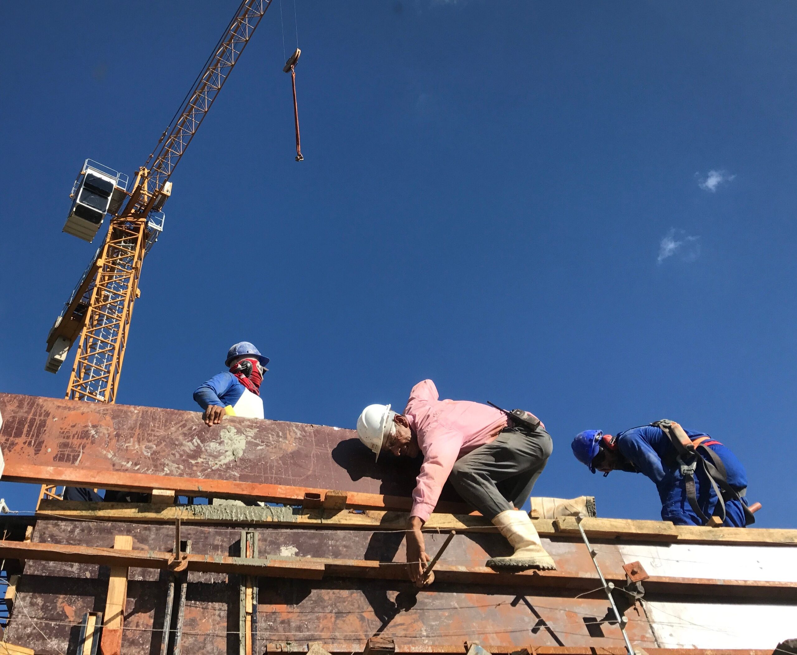 Operários trabalhando em obra de construção civil, com guindaste e céu azul ao fundo