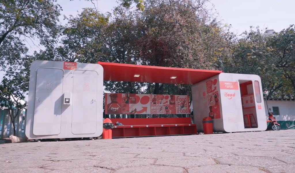 Estrutura modular em vermelho e branco, com banheiros, bancos e cobertura, instalada em uma pra&ccedil;a