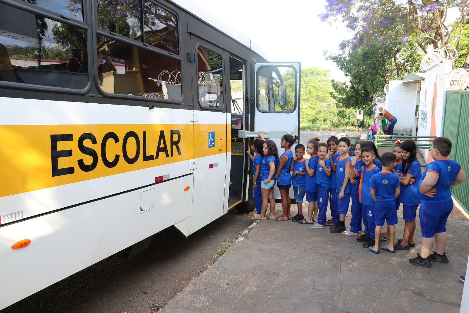 Crianças de uniforme azul aguardam ao lado de ônibus escolar em Goiânia